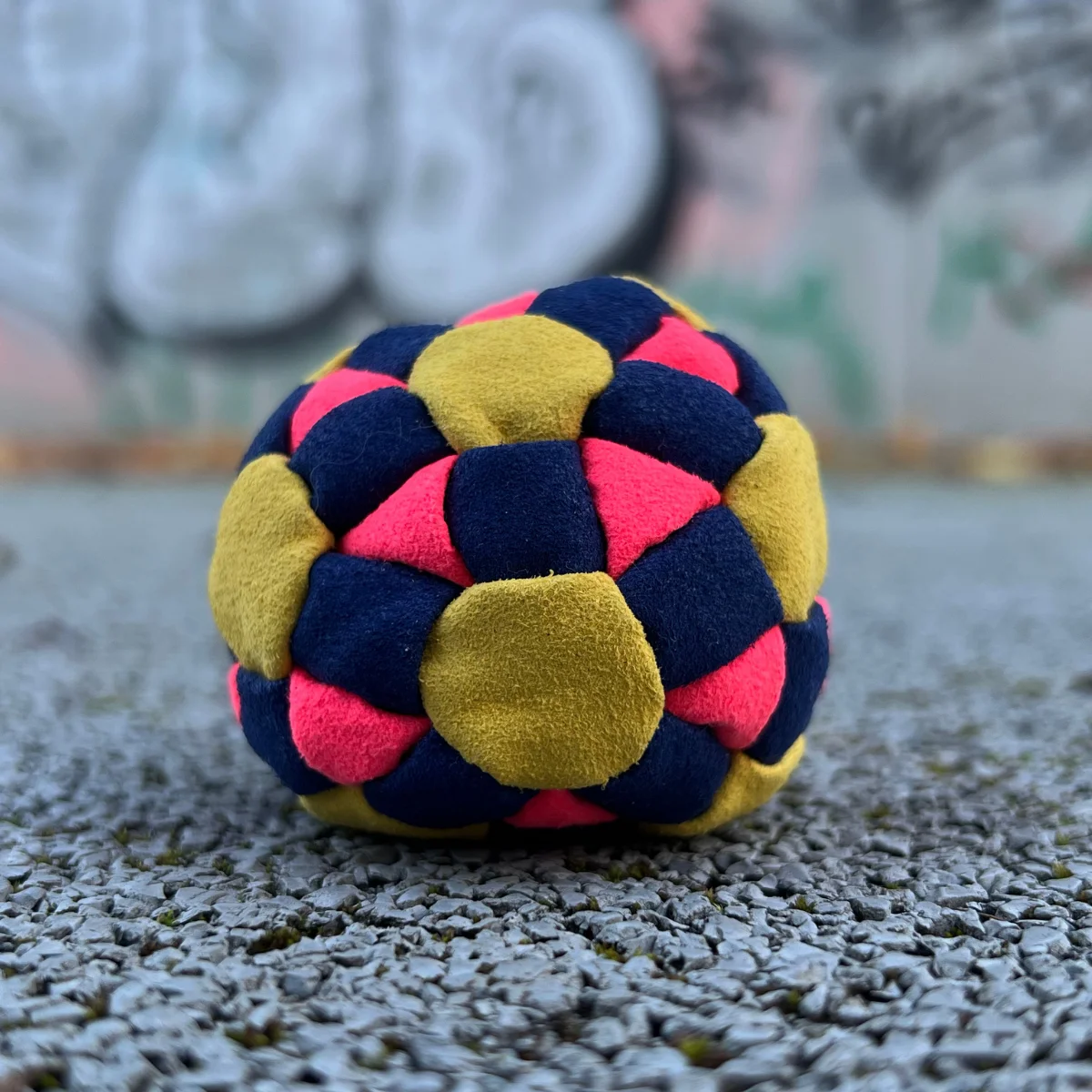 Ein gelb pink dunkelblauer Footbag liegt draußen auf grauem Kies