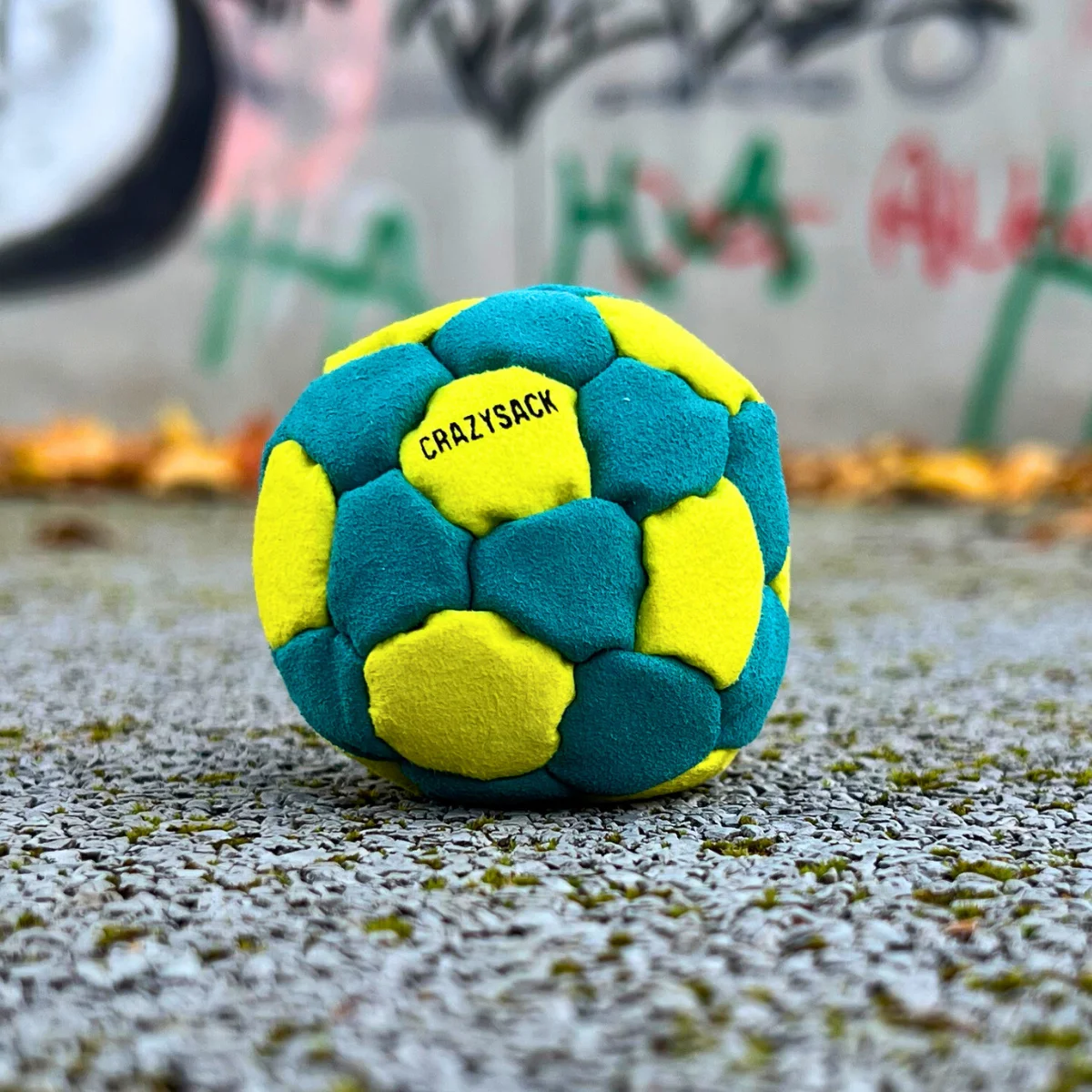Ein hellblau gelber Footbag liegt auf einem grauen Kiesboden im Freien