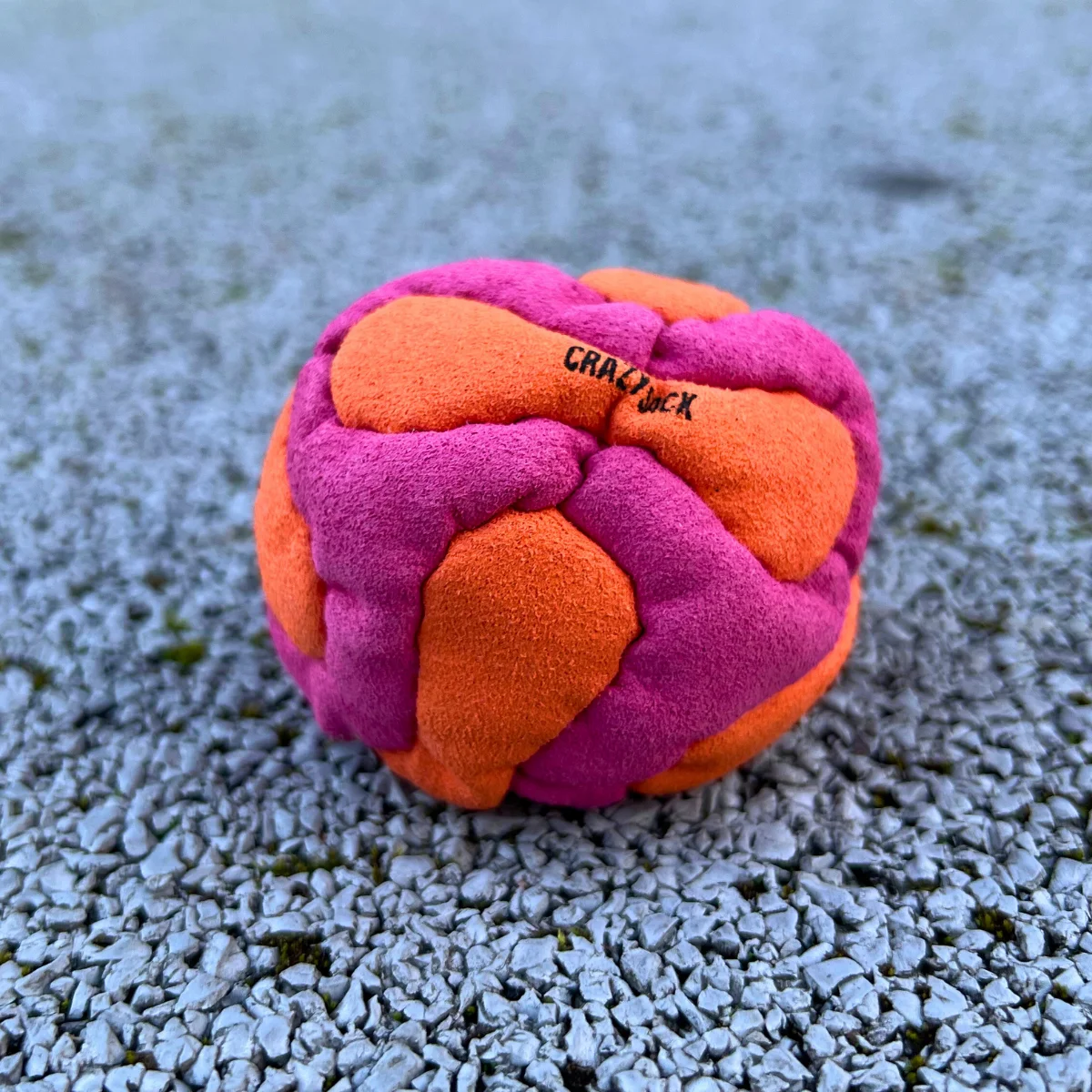 Ein pink orange Footbag liegt auf einem grauen Kiesboden